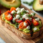 A slice of toasted bread topped with mashed avocado, halved cherry tomatoes, crumbled feta cheese, fresh cilantro, and a drizzle of balsamic glaze. In the background, there are halved avocados and a whole cherry tomato on a wooden cutting board.